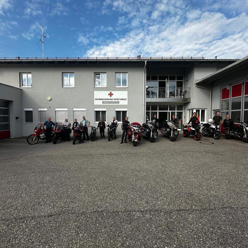 Eine Gruppe von Motorradfahrern steht vor einem Gebäude mit rotem Kreuzzeichen, unter einem teilweise bewölkten Himmel.