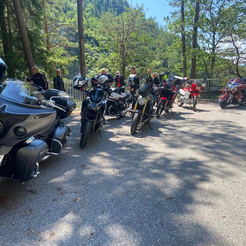 Mehrere Motorräder sind am Straßenrand geparkt, mit Leuten, die herumstehen, in einem Waldgebiet. Helme sind auf den Motorrädern platziert. Das Gebiet ist von Bäumen umgeben und hat einen Holzzaun.