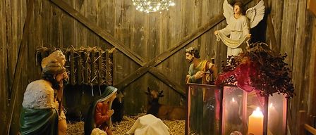 Eine Krippe mit Figuren von Maria, Josef und dem Jesuskind ist in einer Holzscheune ausgestellt, beleuchtet von Weihnachtslichtern und einer Kronleuchter. Ein Engel steht als Wächter über der Szene.