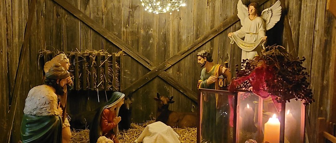 Eine Krippe mit Figuren von Maria, Josef und dem Jesuskind ist in einer Holzscheune ausgestellt, beleuchtet von Weihnachtslichtern und einer Kronleuchter. Ein Engel steht als Wächter über der Szene.