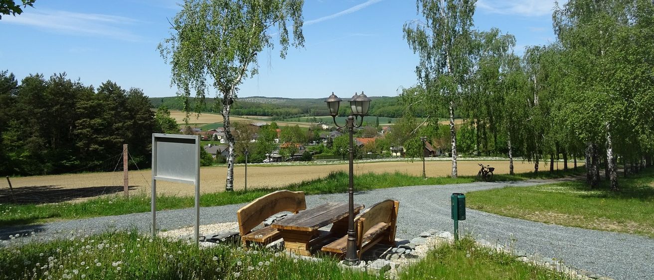 Eine Picknick-Bank mit Holzbänken unter einer Laterne auf einem Kiesweg in einem grasigen Bereich mit Bäumen, einem entfernten Dorf und einem klaren blauen Himmel.