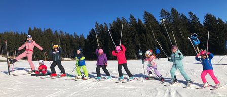 Eine Gruppe von Kindern lernt auf einem verschneiten Hang Skifahren bei blauem Himmel, mit immergrünen Bäumen im Hintergrund.
