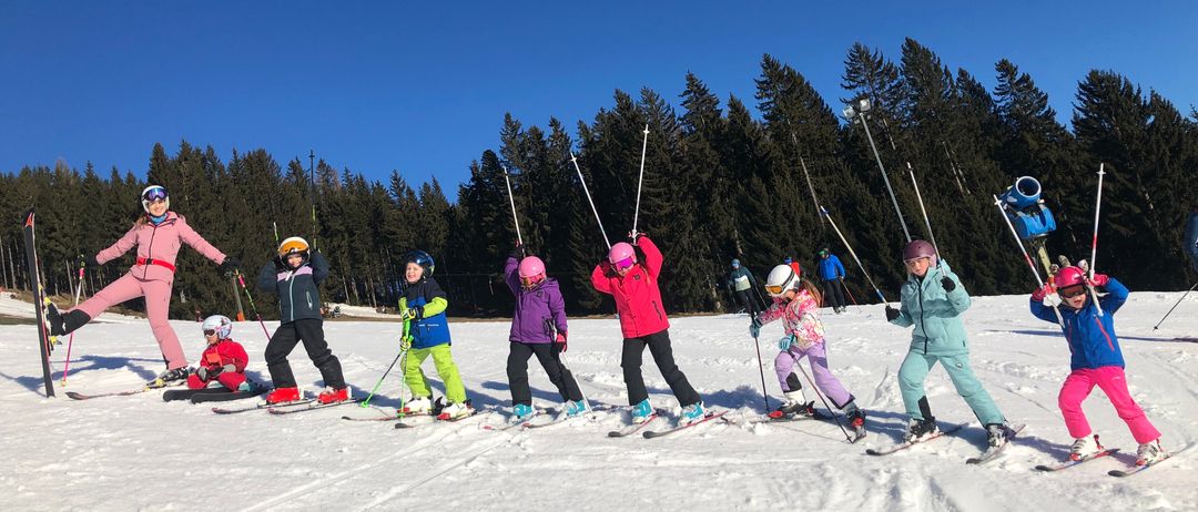 Eine Gruppe von Kindern lernt auf einem verschneiten Hang Skifahren bei blauem Himmel, mit immergrünen Bäumen im Hintergrund.