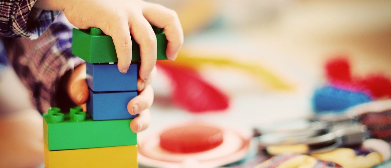 Die Hände eines Kindes bauen einen bunten Stapel aus Bauklötzen. Im Hintergrund sind Spielzeug und Handschuhe zu sehen.