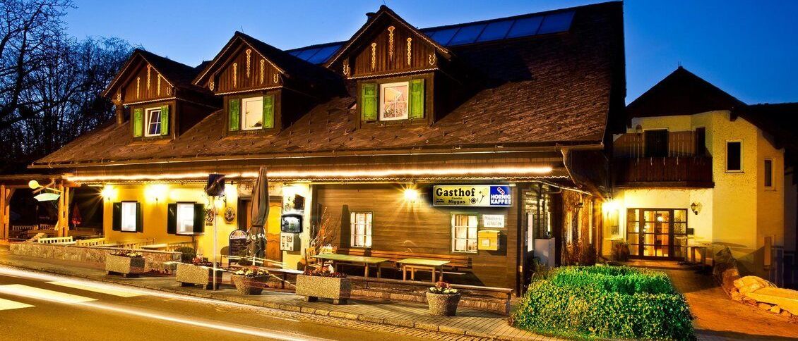 Ein Holzgebäude mit Solarpaneelen auf dem Dach, bei Nacht beleuchtet. Es hat ein Schild mit der Aufschrift 'Gasthof' über dem Eingang.