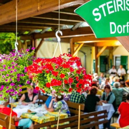 Eine Gruppe von Menschen speisen in einem Restaurant im Freien. Hängende Blumentöpfe mit roten und lila Blumen verleihen Farbe. Ein grünes Schild mit weißen Buchstaben steht für 'Steirisch Dorfw.' Der Hintergrund ist leicht unscharf.