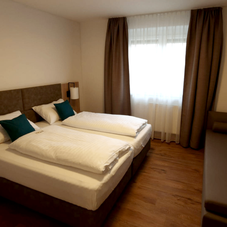 Ein Hotelzimmer mit zwei Betten, weißen Laken und Kissen. Es hat ein Fenster mit weißen Vorhängen und braunen Vorhängen. Das Zimmer hat Holzböden.