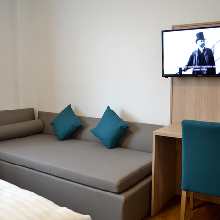 Ein Hotelzimmer mit einem Schlafsofa, zwei blauen Kissen, einem Schreibtisch mit Stuhl und einem Fernseher, der einen Film spielt.