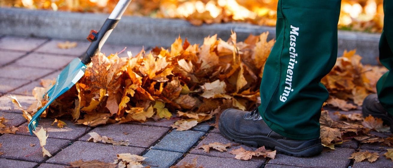 Eine Person in grüner Arbeitskleidung benutzt einen Rechen, um gefallene Blätter auf einem Ziegelweg zu sammeln, mit einem Haufen Blätter im Hintergrund.