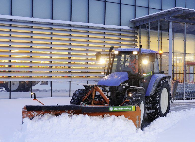 Ein blauer Traktor mit einem Schneepflug räumt Schnee vor einem Gebäude mit Glaswänden.