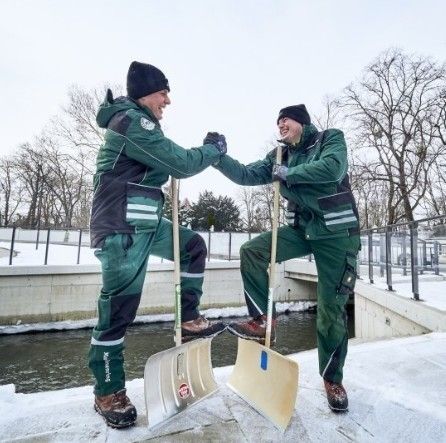 Zwei Männer in grünen Uniformen stehen auf einer Brücke, halten Schneeschaufeln und schütteln Hände. Sie sind in der Nähe eines kleinen Baches, und das Gebiet ist schneebedeckt.