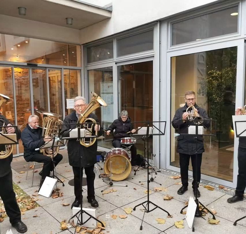 A group of musicians are playing instruments outside a building. They are playing trumpets, tubas, and drums. The building has glass walls and doors.