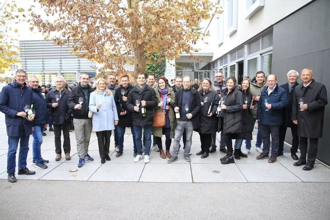 A group of people stands outside in winter clothing, holding wine glasses and bottles, with a tree and building in the background.