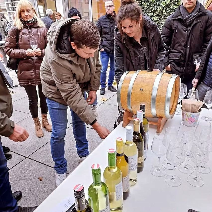 A group of people are gathered around a table with wine bottles and glasses. A man is pouring wine from a barrel.