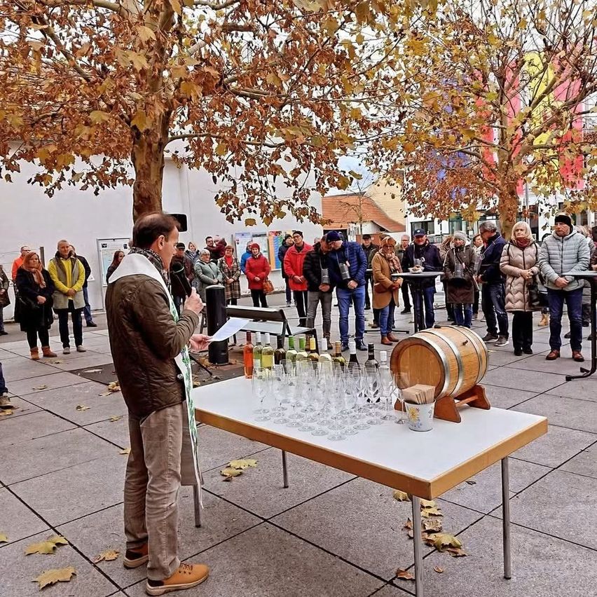 A man speaks to a crowd around a table with wine glasses and a barrel, standing among trees with fallen leaves.