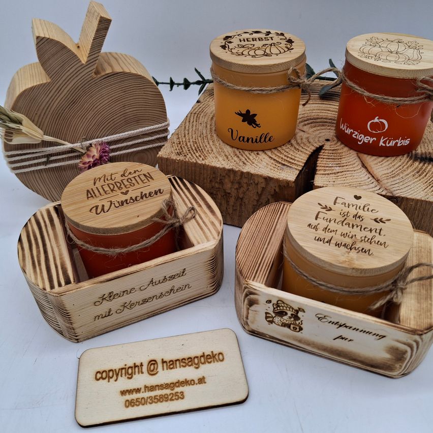 Eine Sammlung von Holzgläsern mit personalisierten Deckeln, ausgestellt auf Holzstämmen. Jedes Glas hat ein einzigartiges Etikett mit Text wie 'Vanille' und 'Würziger Kürbis'. Ein Holzapfel und eine kleine Karte mit einer Website und Telefonnummer sind ebenfalls vorhanden.