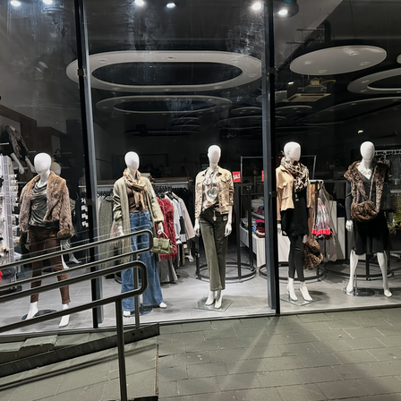 Ein Schaufenster mit fünf Mannequins, die in verschiedenen Kleidungsstücken wie Jacken, Schals und Hosen gekleidet sind, mit einem Metallgeländer auf der linken Seite.