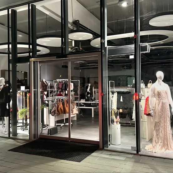 Ein modernes Bekleidungsgeschäft mit Glaswänden, das Mannequins in verschiedenen Outfits und ausgestellten Taschen zeigt. Der Eingang ist offen, und es gibt runde Lampen an der Decke.