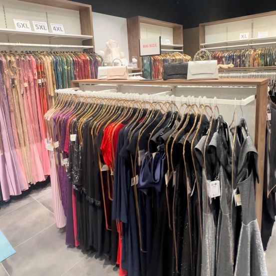 Ein Bekleidungsgeschäft zeigt eine Vielzahl von Kleidern an Kleiderbügeln, mit Größenetiketten wie 6XL, 5XL und 4XL. Die Kleider sind in verschiedenen Farben und Stilen, einige mit Pailletten und andere schlicht.