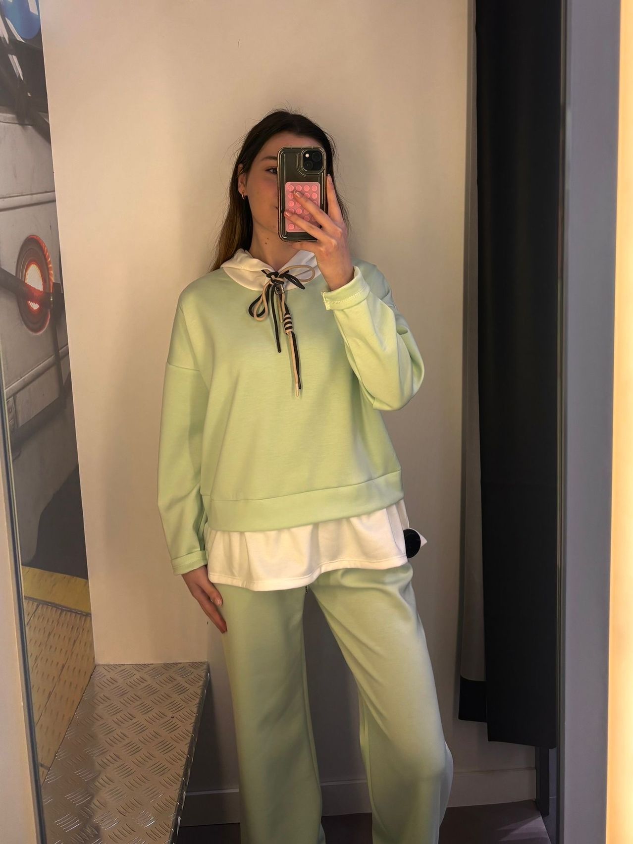A woman takes a selfie with a cellphone. She wears a mint-green sweatshirt and pants. The background is a white wall with a mirror.
