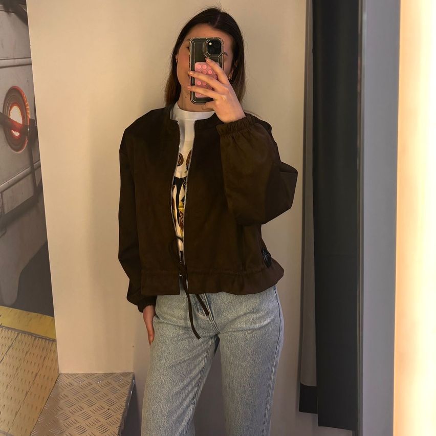 A woman takes a selfie in front of a mirror wearing a brown suede jacket and light blue jeans.