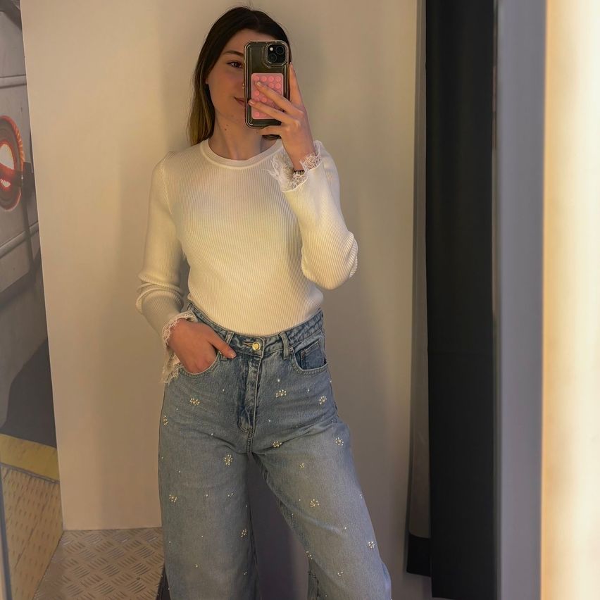 A woman takes a selfie with a phone. She is wearing a white top and embellished jeans. She stands against a light-colored wall.