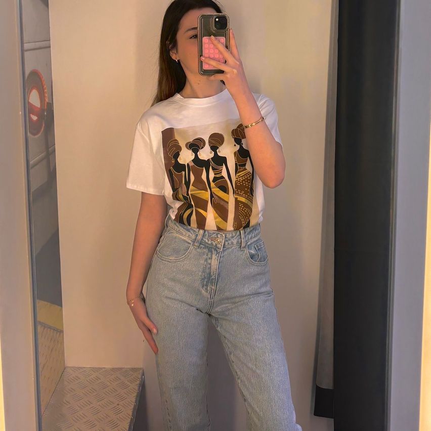 A woman in a room takes a selfie wearing a white t-shirt with a graphic design, blue jeans, and a bracelet.
