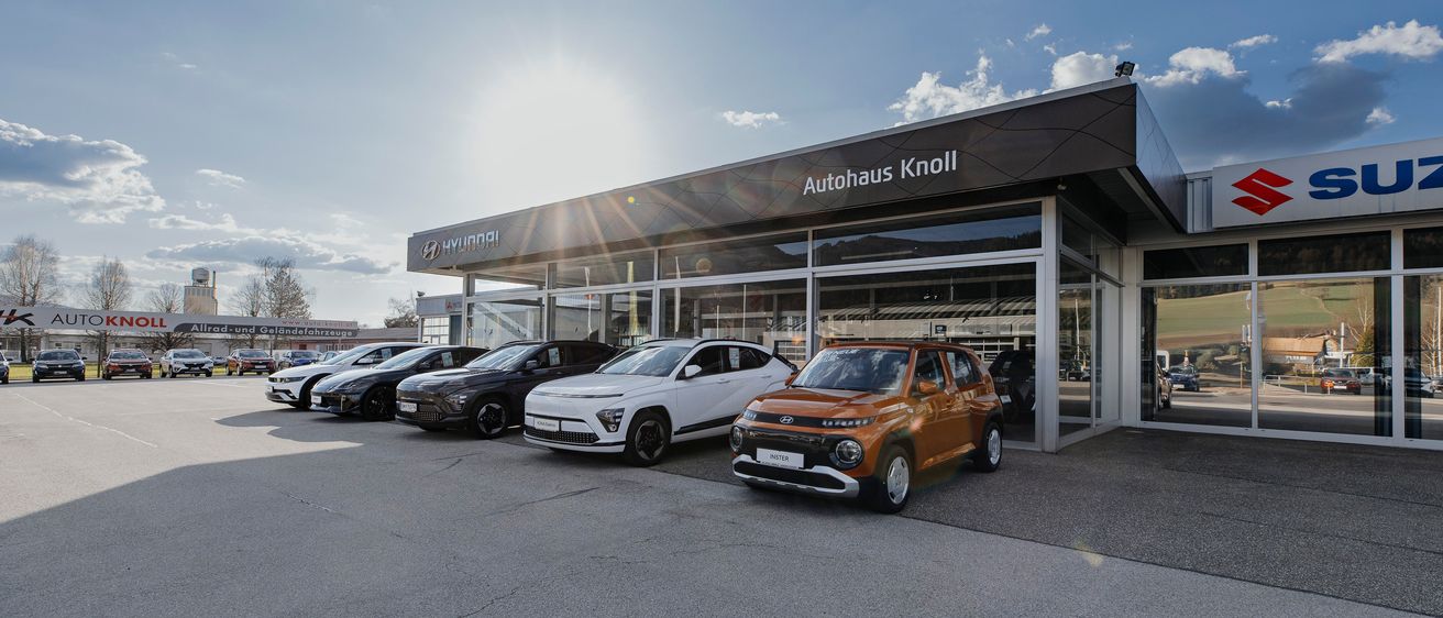 Das Hyundai Knoll-Autohaus zeigt eine Auswahl an Autos unter einem strahlenden Himmel. Das Schaufenster hat ein modernes Design mit großen Glasfenstern und einem prominenten Schild, das 'Autohaus Knoll' steht. Mehrere Autos, darunter ein orangefarbener SUV, sind vor dem Gebäude geparkt.