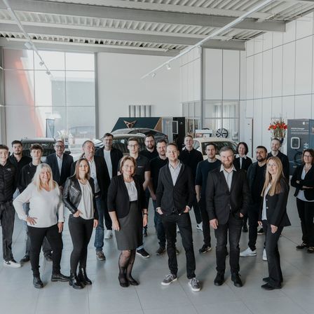 Eine Gruppe von Personen in formeller Kleidung posiert für ein Foto in einer Ausstellungshalle mit ausgestellten Autos im Hintergrund.