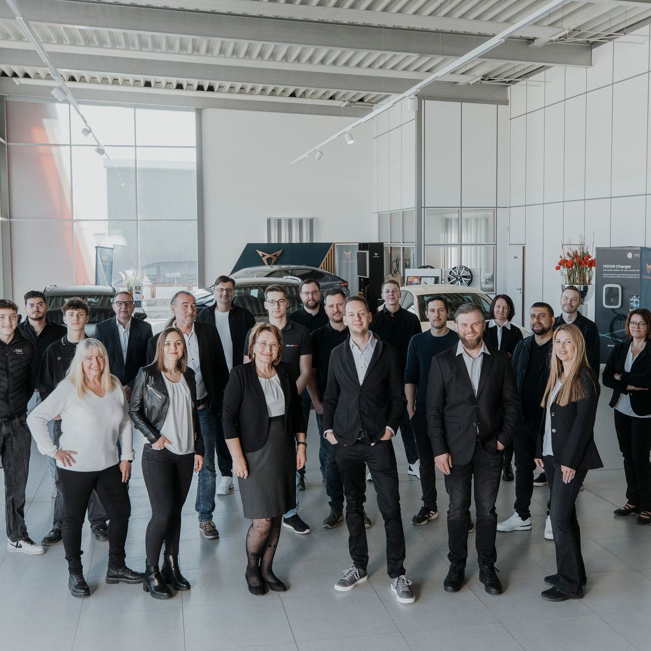 Eine Gruppe von Personen in formeller Kleidung posiert für ein Foto in einer Ausstellungshalle mit ausgestellten Autos im Hintergrund.