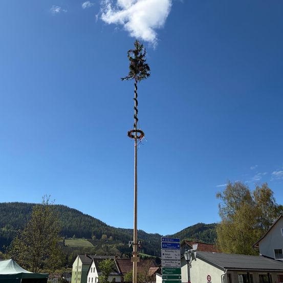 Ein hoher Baum ist an einem Pfosten mit einem Kranz und einer kleinen Flagge in einem Dorf mit Bergen und Häusern im Hintergrund befestigt.