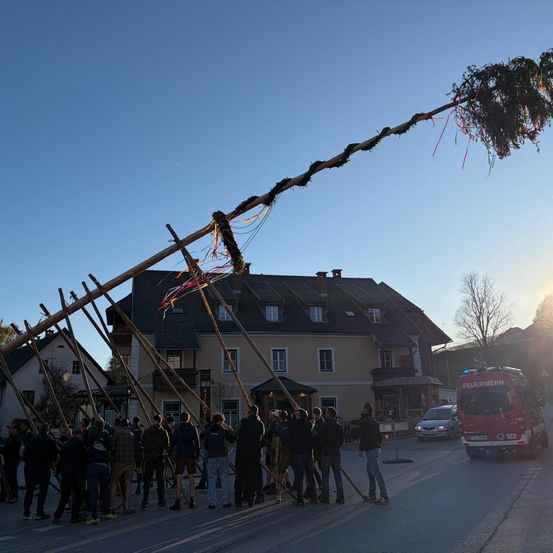 Eine Menschenmenge versammelt sich um eine geschmückte Holzstruktur, möglicherweise ein Maibaum, in der Nähe von Häusern. Ein Feuerwehrauto steht am Straßenrand.