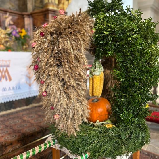 Eine dekorative Weizengarbe mit rosa Blumen und einer Kürbis, umgeben von grünem Laub, auf einem Tisch in einer Kirche ausgestellt.