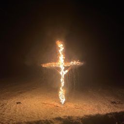 Ein brennendes Kreuz in der dunklen Nacht sticht im schneebedeckten Boden hervor. Flammen beleuchten das Kreuz am Nachthimmel.