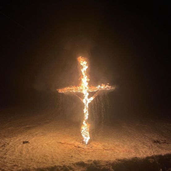 Ein brennendes Kreuz in der dunklen Nacht sticht im schneebedeckten Boden hervor. Flammen beleuchten das Kreuz am Nachthimmel.