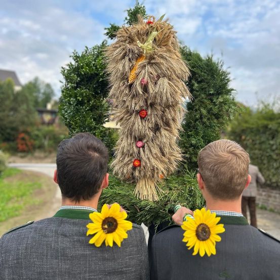Zwei Männer mit Sonnenblumen auf dem Rücken stehen vor einem großen Kranz, wahrscheinlich bei einem Fest.