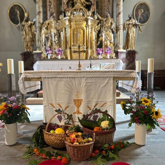 Ein Kirchenaltar ist mit einem Kreuz geschmückt, umgeben von Körben mit Obst und Gemüse. Er hat ein weißes Tuch mit einem goldenen Kelchsymbol. Zwei brennende Kerzen und Blumen befinden sich auf beiden Seiten. Dahinter stehen Statuen und ein vergoldeter Altar.