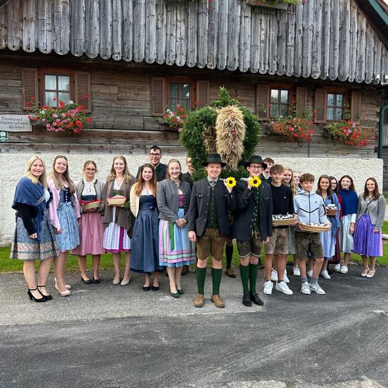 Eine Gruppe von Menschen in traditioneller bayerischer Kleidung posiert für ein Foto vor einem rustikalen Gebäude mit Blumen. Die beiden Männer in der Mitte halten Sonnenblumen.
