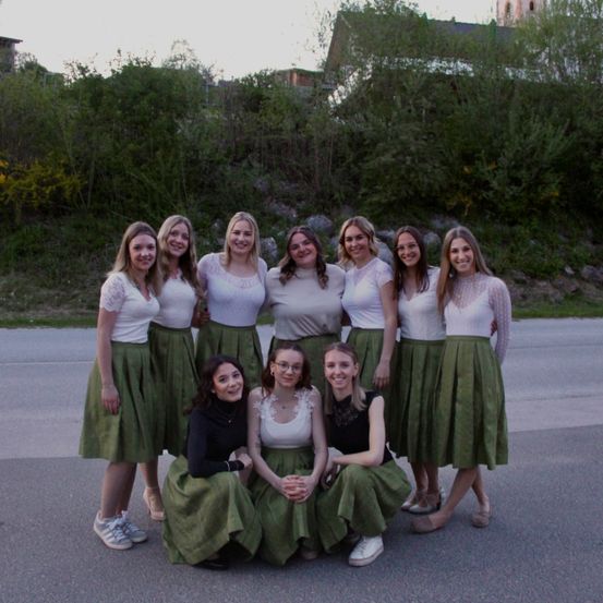 Neun Frauen in weißen Oberteilen und grünen Röcken posieren für ein Foto auf einer Straße, mit Grün und einem Gebäude im Hintergrund.