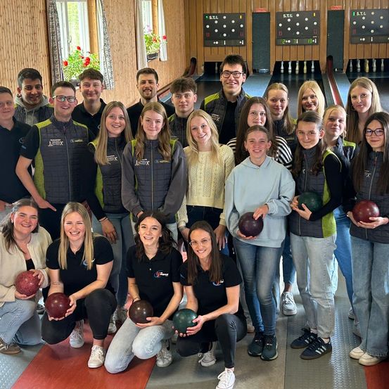 Eine Gruppe von Menschen posiert für ein Foto in einer Bowlingbahn. Einige halten Bowlingkugeln und lächeln. Hinter ihnen befinden sich Bowlingbahnen mit Anzeigetafeln.