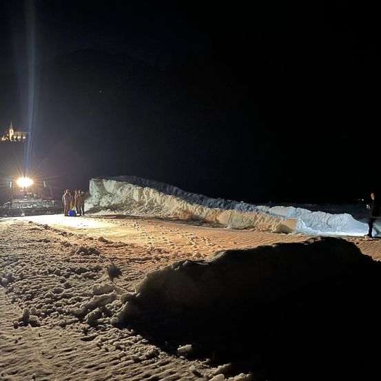 Eine Gruppe von Menschen steht bei Nacht im Schnee mit einem Fahrzeug in der Nähe. Ein Licht scheint in der Ferne.