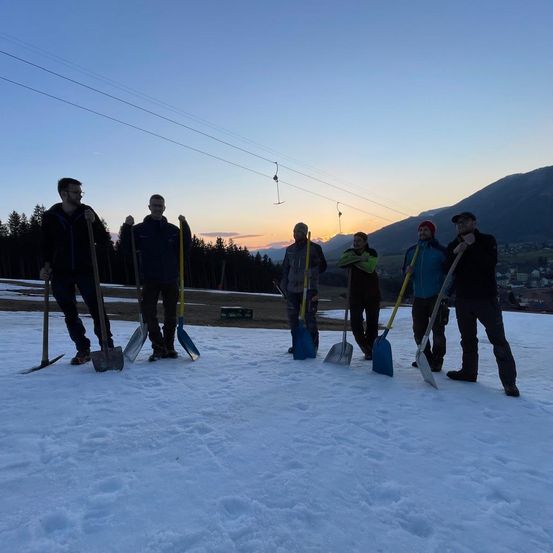 Eine Gruppe von Männern, die Schnee schaufeln, mit einem Sonnenuntergang im Hintergrund und Bergen in der Ferne.
