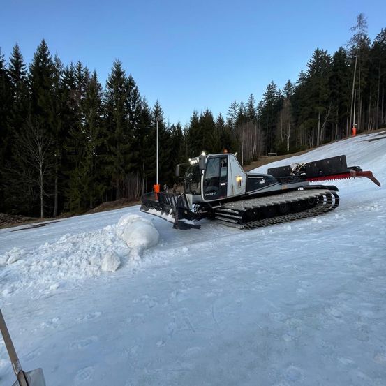 Eine Schneefräse steht auf einem verschneiten Hang. Bäume sind im Hintergrund, und ein klarer blauer Himmel ist oben.