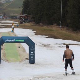 Ein Skifahrer fährt in einem von Bäumen und Bergen umgebenen Gebiet. Ein blauer Bogen zeigt den Text Diepold und es stehen mehrere Personen herum.