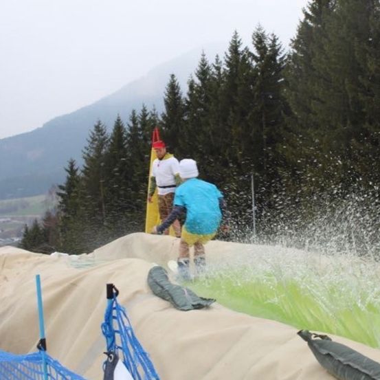 Zwei Personen befinden sich auf einer Wasserrutsche, eine in einem blauen Outfit und die andere in Gelb. Bäume und Berge sind im Hintergrund.