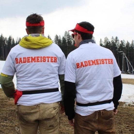 Zwei Männer, die weiße Jacken mit 'Bademeister' tragen, stehen auf einem verschneiten Feld, möglicherweise in einem Skigebiet, mit Bäumen und Bergen im Hintergrund.