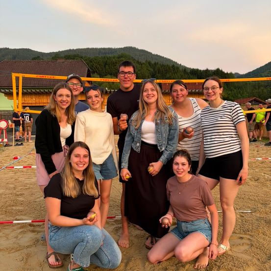 Eine Gruppe von Menschen posiert für ein Foto in einem sandigen Bereich mit einem Volleyballnetz im Hintergrund. Sie lächeln und halten Tennisbälle.