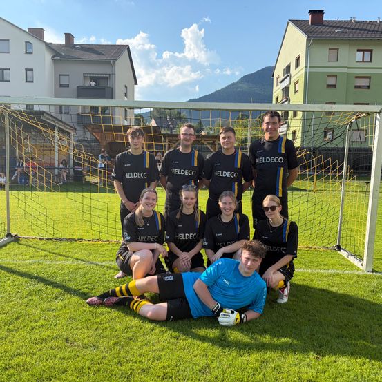 Eine Fußballmannschaft posiert für ein Foto vor einem Tor. Sie tragen einheitliche Trikots mit 'HELDECO' bedruckt. Im Hintergrund sind Gebäude und ein Berg zu sehen.