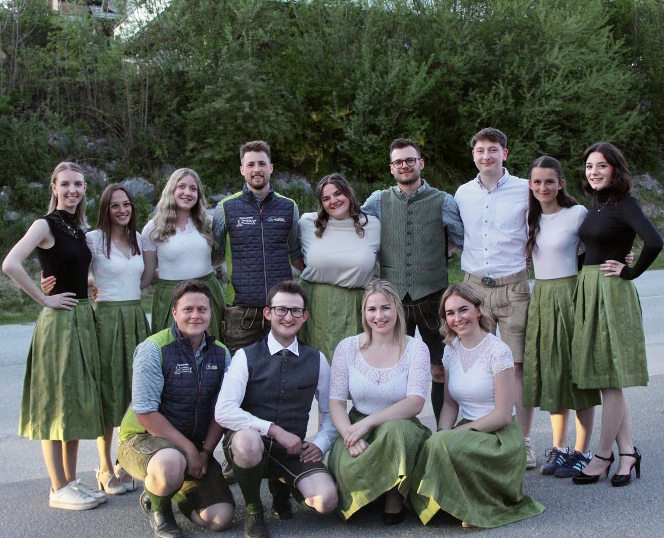 Eine Gruppe von Menschen posiert für ein Foto in einem Außenbereich mit Bäumen im Hintergrund. Sie lächeln alle und tragen traditionelle deutsche Kleidung.