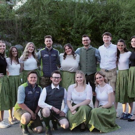 Eine Gruppe von Menschen posiert für ein Foto in einem Außenbereich mit Bäumen im Hintergrund. Sie lächeln alle und tragen traditionelle deutsche Kleidung.